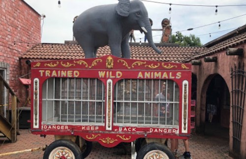 A circus-style cart with a statue of an elephant on top, labeled "Trained Wild Animals," in a playful outdoor setting.