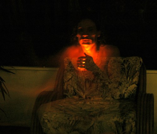 A person sits on a patterned chair in dim light, holding a glowing object, creating an eerie atmosphere.