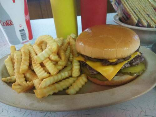 A cheeseburger with pickles and crinkle-cut fries on a plate, with ketchup and mustard bottles in the background.