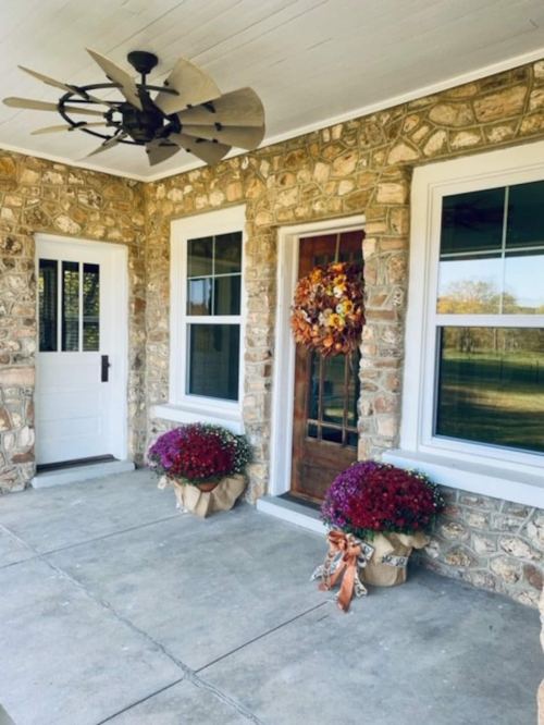 A cozy porch with stone walls, a wooden door, a wreath, and potted mums in decorative bags.