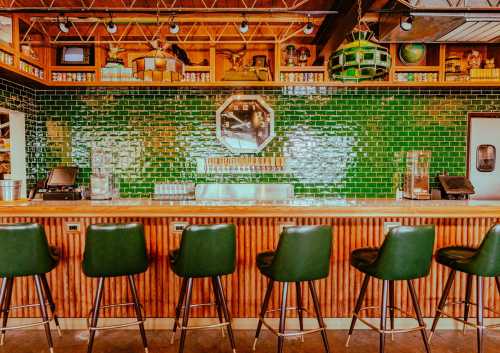 A stylish bar with a green tiled backsplash, wooden accents, and high stools, creating a vibrant and inviting atmosphere.