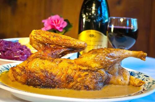A roasted chicken on a plate with gravy, accompanied by red cabbage, wine, and a flower in the background.