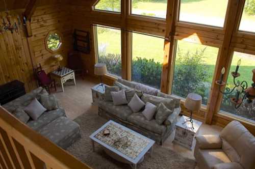 Cozy wooden living room with large windows, comfortable sofas, and a coffee table, surrounded by greenery outside.