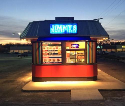 A brightly lit fast-food stand named "Jimmy's" at dusk, featuring a colorful menu and drive-thru window.