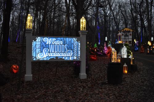 A sign reading "Winter Woods Spectacular" surrounded by colorful holiday lights in a wooded area.