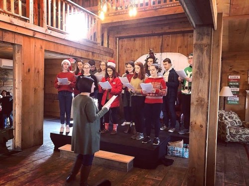 A choir in festive attire performs on a stage in a rustic wooden venue, with a conductor leading them.