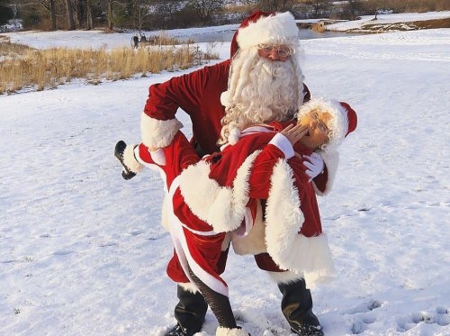 A man in a Santa costume lifts a woman in a Santa outfit, both smiling in a snowy landscape.