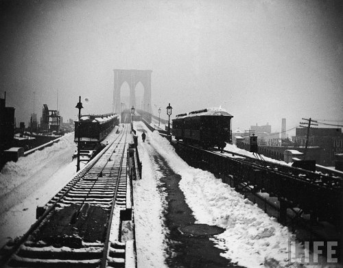 A snowy scene of the Brooklyn Bridge with train tracks and a lone figure walking in the distance.