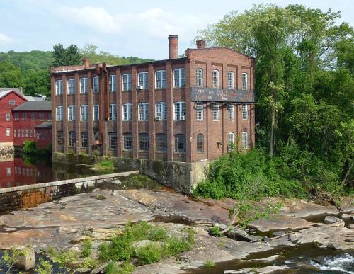 Historic brick mill building by a river, surrounded by greenery and rocky terrain.
