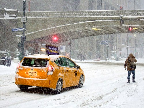 A yellow taxi drives through a snowy street while a person walks nearby, with heavy snowfall and traffic lights visible.