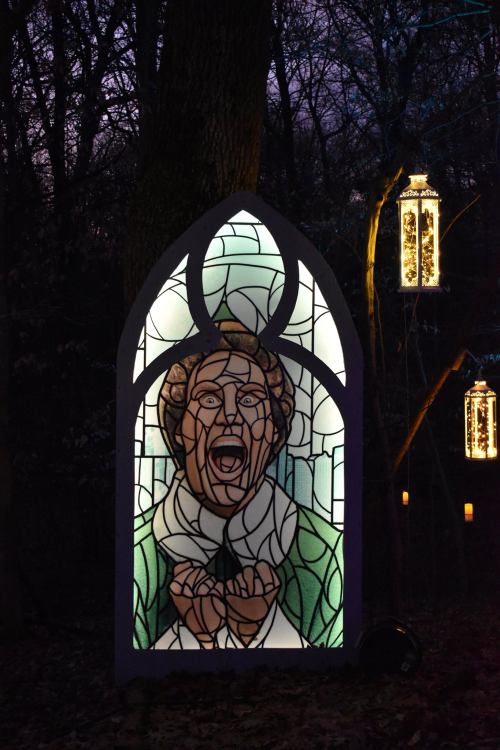 A stained glass window depicting a figure with an expressive face, surrounded by trees and lanterns at dusk.