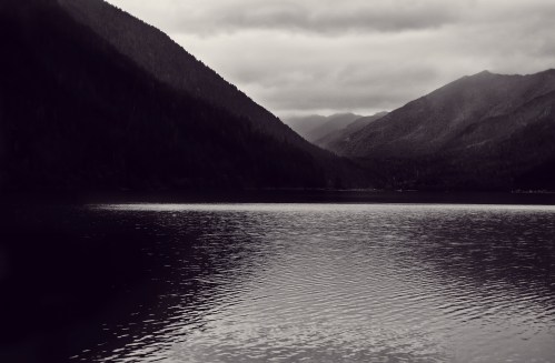 A serene lake surrounded by dark mountains under a cloudy sky, captured in black and white.