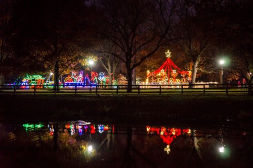 Colorful holiday lights illuminate a park at night, reflecting in a calm pond surrounded by trees.