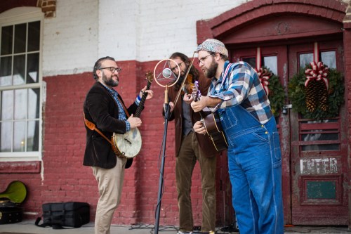 Three musicians perform outdoors, playing a banjo and guitar, with a vintage microphone and festive decorations in the background.