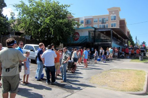 A long line of people waiting outside a building on a sunny day, with trees and a hotel in the background.