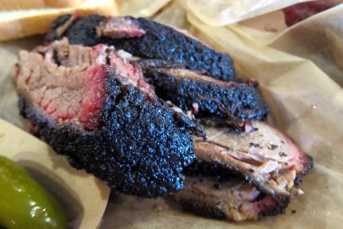 Sliced smoked brisket with a dark, charred crust, served on parchment paper with pickles and bread.