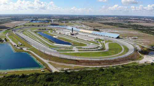 Aerial view of a large racetrack surrounded by water bodies and green fields under a blue sky with scattered clouds.