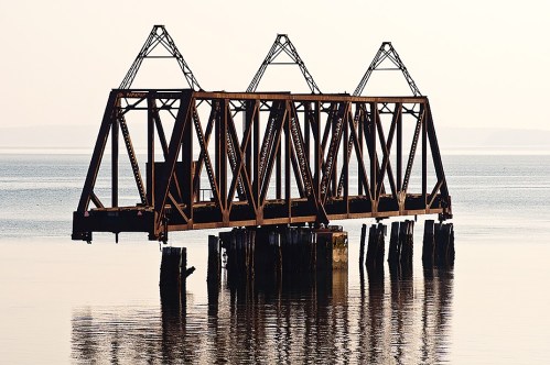 Rusty train trestle bridge stands over calm water, reflecting its structure in the serene landscape.