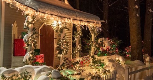 A cozy, snow-covered house adorned with festive lights and decorations, surrounded by wrapped gifts and greenery.
