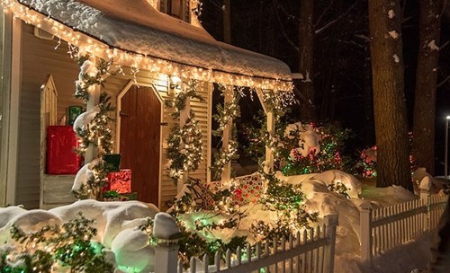 A cozy, snow-covered house decorated with festive lights and greenery, surrounded by a white picket fence.