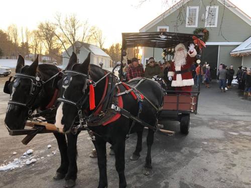 A horse-drawn wagon with Santa waving, surrounded by people and festive decorations in a winter setting.