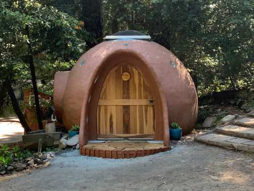 A round, clay-colored structure with a wooden door, surrounded by greenery and a gravel path.