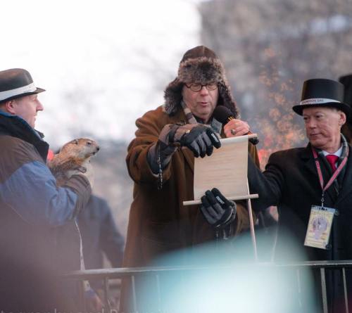 A man in a hat holds a groundhog while another man reads a sign, both on a stage during a winter event.