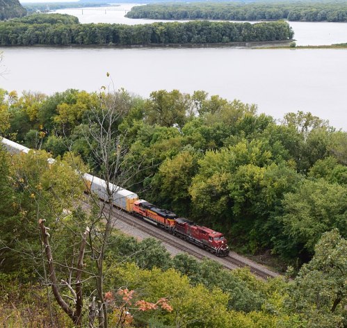 A train travels along a riverbank, surrounded by lush greenery and rolling hills under a cloudy sky.
