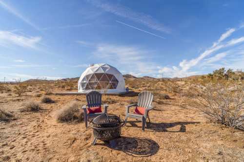 A geodesic dome in a desert landscape, with two chairs and a fire pit in the foreground under a clear blue sky.