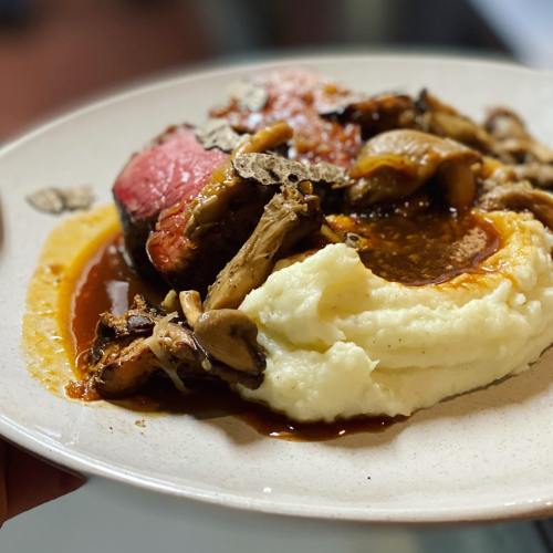 A plate of sliced beef with mushrooms and gravy, served alongside creamy mashed potatoes.