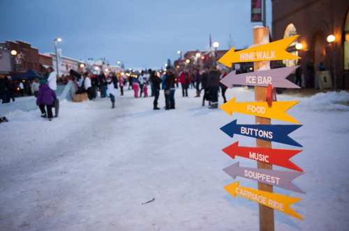 A snowy street scene with people enjoying a winter festival, featuring a signpost with colorful directional arrows.