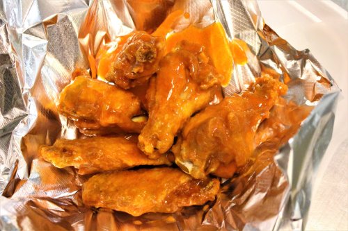 A serving of crispy chicken wings coated in a spicy orange sauce, placed in a foil-lined container.