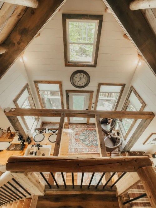 A cozy cabin interior viewed from above, featuring wooden beams, large windows, and a decorative rug.