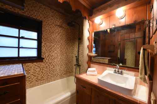 A cozy bathroom featuring a bathtub, sink, wooden walls, and a large mirror with warm lighting.