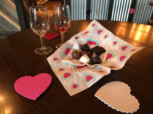 Two wine glasses beside a plate of assorted chocolates on a heart-patterned napkin, with heart-shaped coasters.