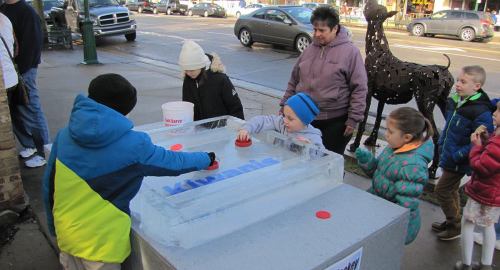 Children playing a game on an ice block outside, with adults observing and a metal sculpture in the background.