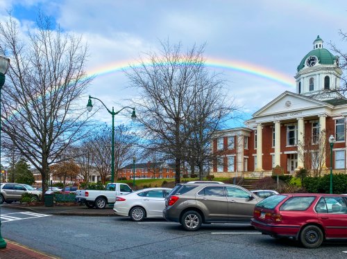 A rainbow arcs over a historic building, with parked cars and bare trees in a town square setting.