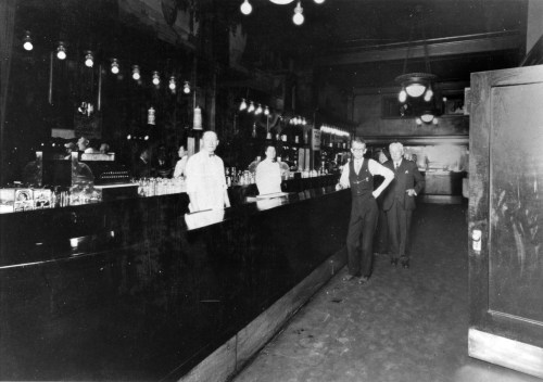 A vintage bar scene with a long counter, bartenders, and patrons in early 20th-century attire.