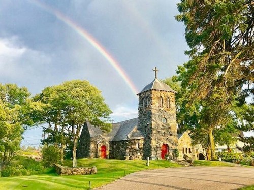 A stone church with a rainbow overhead, surrounded by green trees and a grassy landscape.