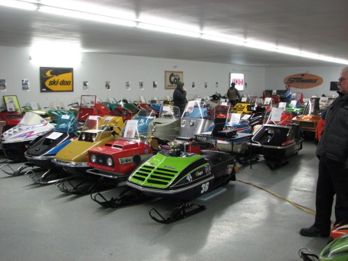 A collection of vintage snowmobiles displayed in a spacious indoor setting, with visitors observing the exhibits.