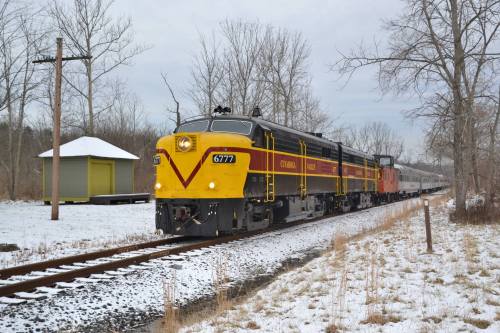 A vintage train with a yellow and red locomotive travels along snowy tracks near a small station and bare trees.