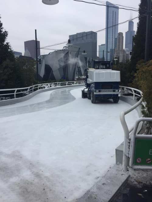 A zamboni cleans an ice rink with a city skyline in the background on a cloudy day.