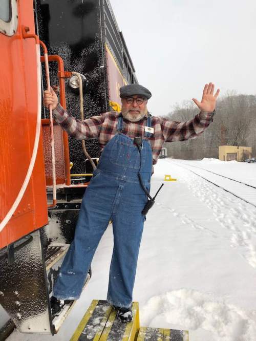 A man in overalls and a cap stands on a train step, waving in a snowy landscape.