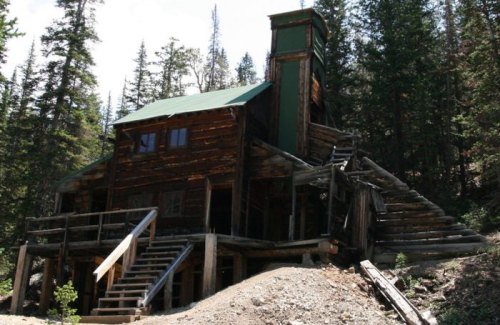 A rustic wooden building with a green roof, surrounded by trees and a dirt path leading to its stairs.