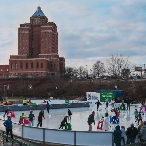 A bustling outdoor ice skating rink with a historic building in the background and people skating joyfully.