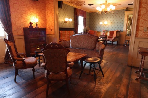 Cozy vintage room with ornate furniture, a wooden table, and warm lighting, featuring patterned wallpaper and wooden floors.