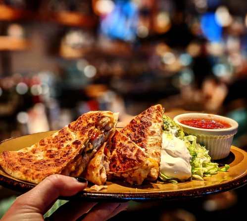 A hand holding a plate with three quesadilla wedges, lettuce, sour cream, and a small bowl of salsa.