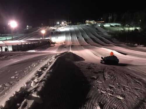 A snowy tubing hill at night, with lights illuminating the slopes and a person sliding down in the foreground.