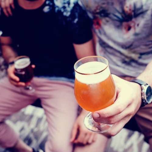 A close-up of a hand holding a glass of beer, with two friends in the background enjoying drinks.