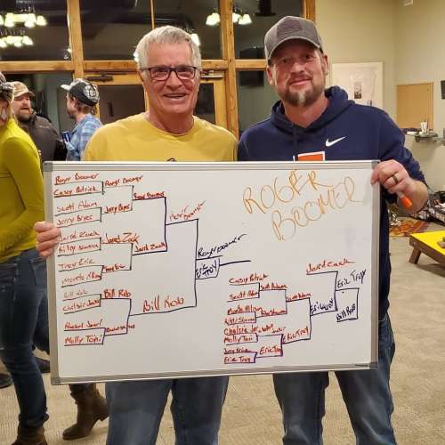 Two men stand beside a whiteboard displaying a tournament bracket, with names and match results written on it.